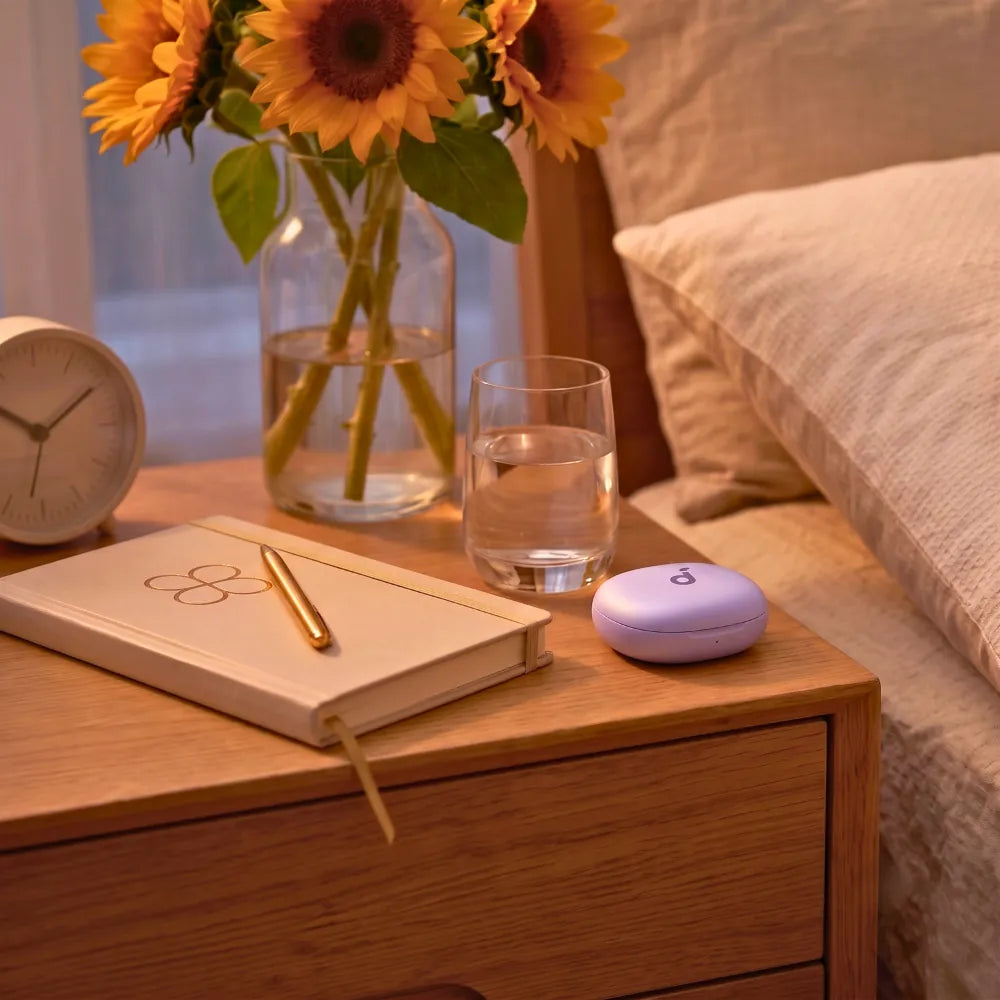 A white sleepbuds charging case sits on a wooden bedside table next to a glass of water, notebook, pen, clock, and a vase of sunflowers in a bedroom setting.